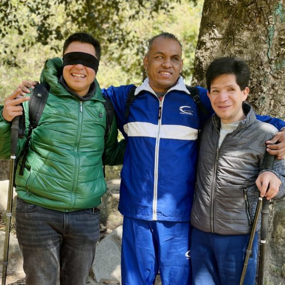 Fotografía de tres turistas en el bosque de los dinamos. Dos tienen discapacidad visual y el tercero los ojos vendados. Los tres están abrigados con chamarras y portan bastones de trekking.