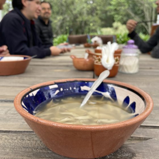Fotografía de una sopa de hongos en un plato de barro sobre una mesa de madera alargada, del restaurante de la cima de la montaña. Al fondo se ven cinco personas platicando y jarritos de barro con agua de sabor.